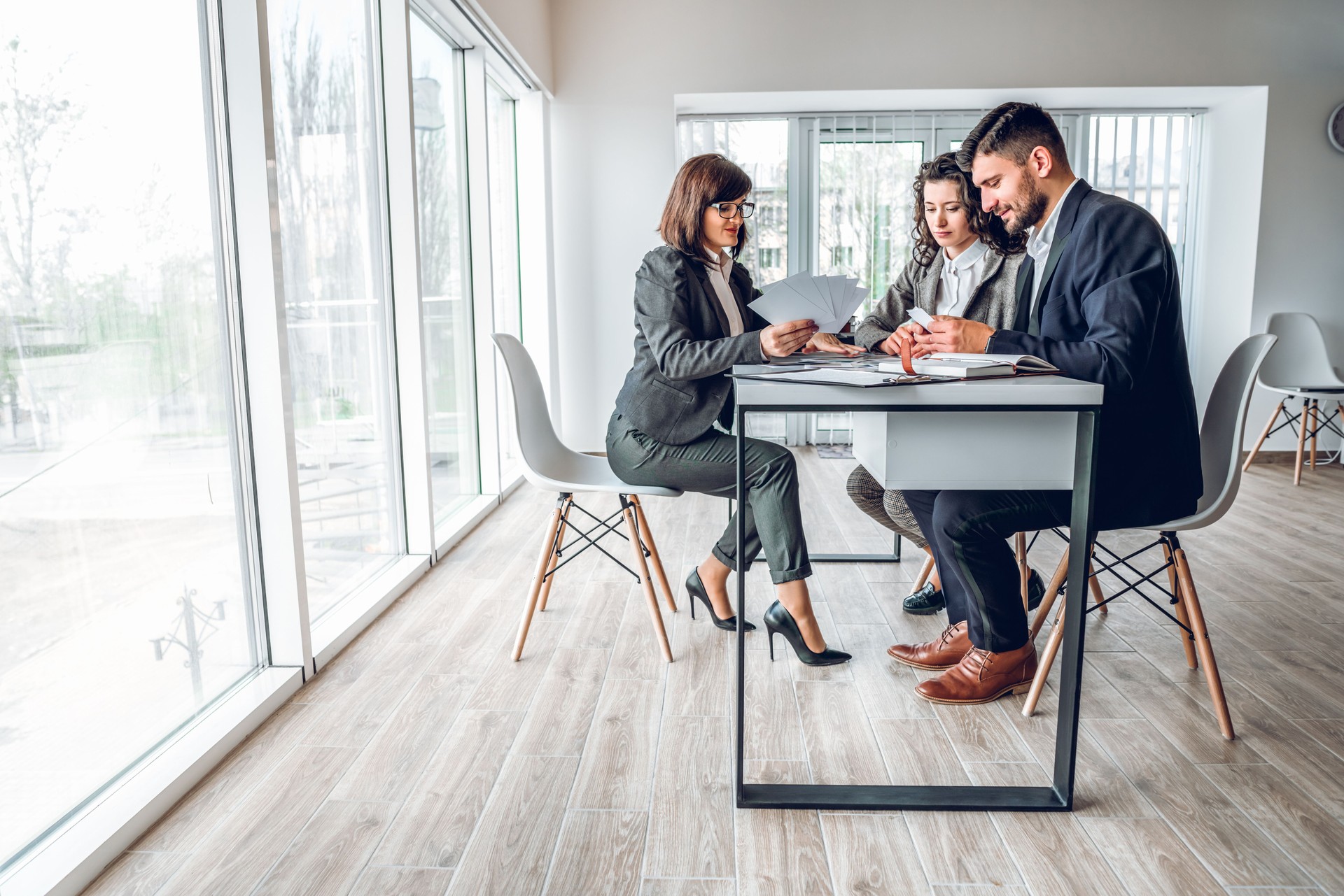Wide angle view of group business people in bright spacious modern office near panoramic windows. Young businessman and businesswoman sitting at the desk in design studio with mature woman sales manager. Work with client concept