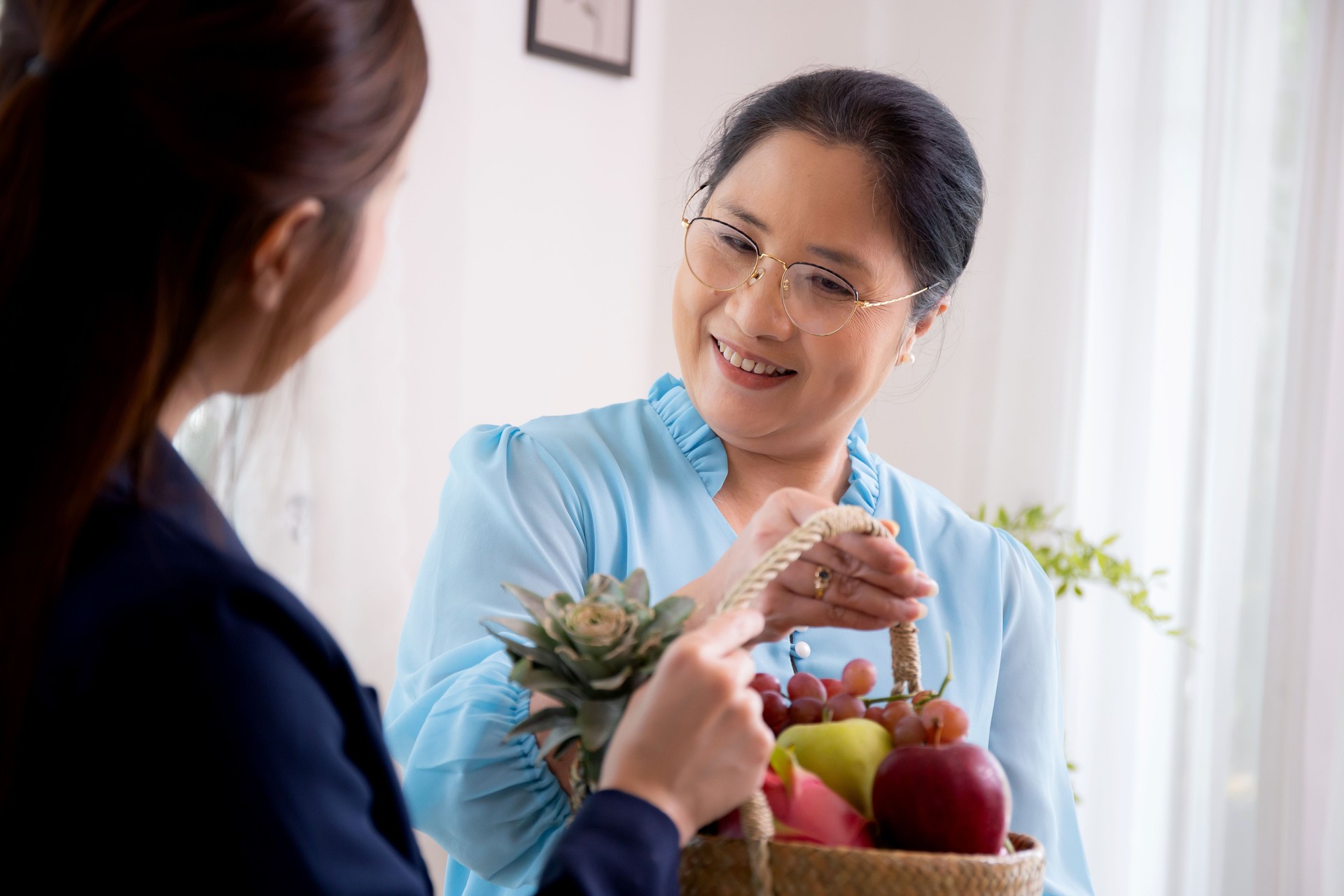 Young asian woman agent insurance visit customer elderly while giving fruit basket in the living room at home, businesswoman or consultant talking and service client with senior, business concept.